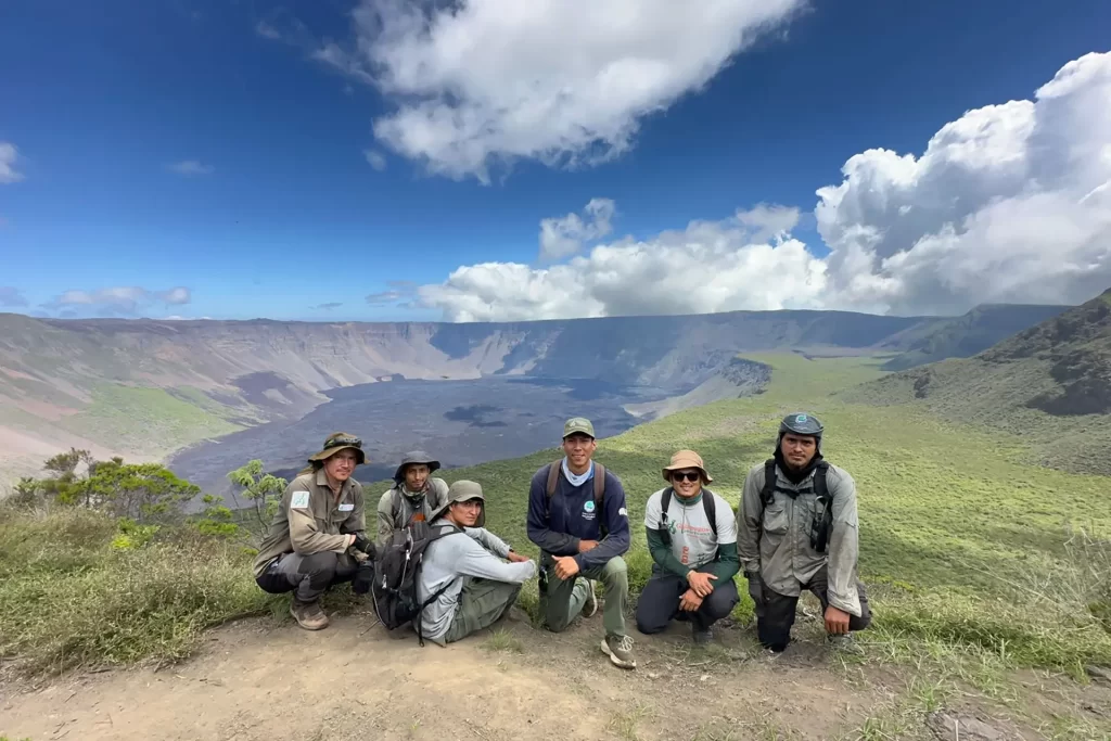 The conservation expedition team, from left to right: Jorge Carrión, Adrián Cueva, Danny García, Sebastián Ballesteros, Ronny Gil, and Paúl Vaca, stands at the summit of Wolf Volcano, with the neighboring island of Fernandina in the background, where the Cumbre volcano is currently erupting.